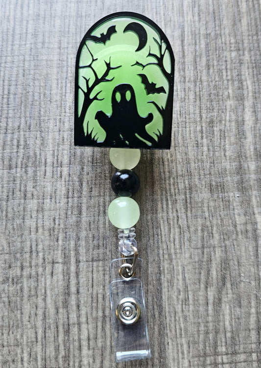Green glow-in-the-dark tombstone-shaped badge reel with an eerie ghost emerging from the ground, surrounded by trees, a moon, and bats, with matching glow-in-the-dark decorative beads.