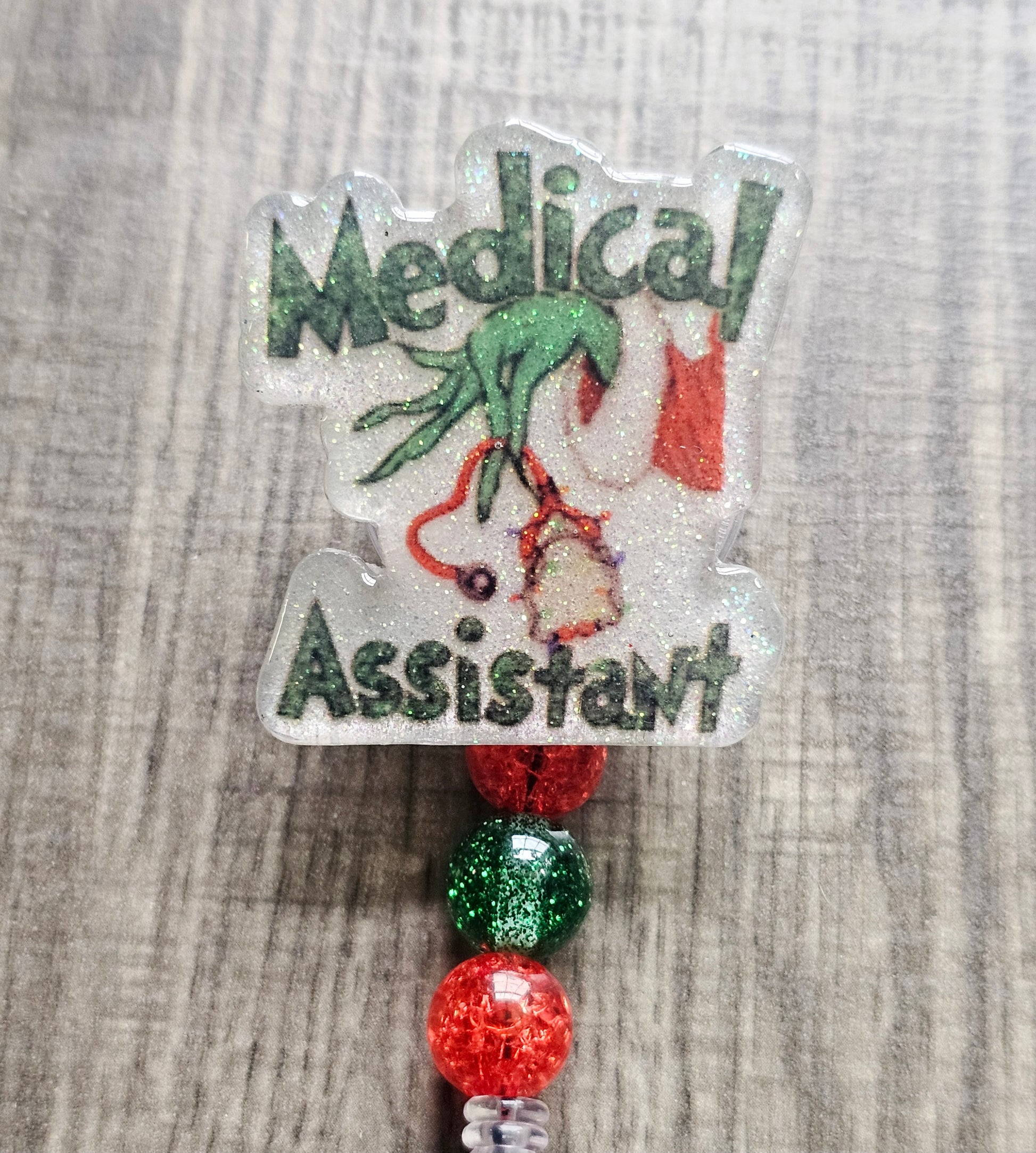 Badge reel depicting the Grinch's hand in a festive sweater holding stethoscopes, with the words "Medical Assistant" in a Christmas theme.