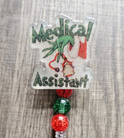 Badge reel depicting the Grinch's hand in a festive sweater holding stethoscopes, with the words "Medical Assistant" in a Christmas theme.