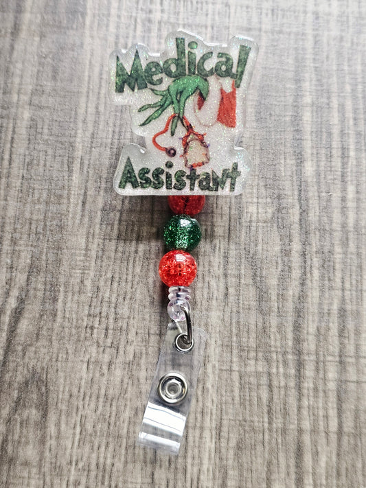 Badge reel depicting the Grinch's hand in a festive sweater holding stethoscopes, with the words "Medical Assistant" in a Christmas theme.