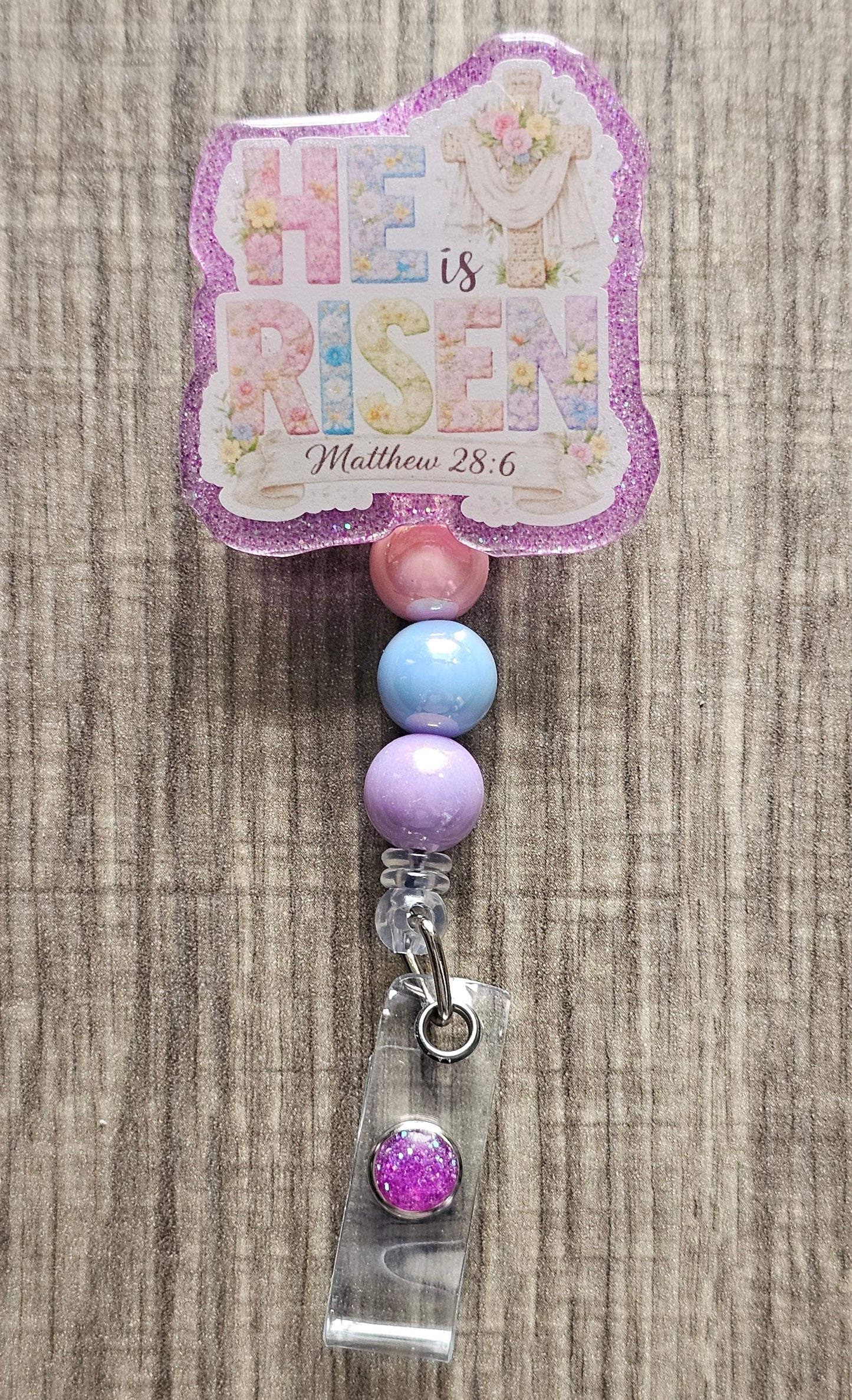 Retractable badge reel featuring "He Is Risen" floral typography with a cross, purple robe, and Matthew 28:6 scripture.