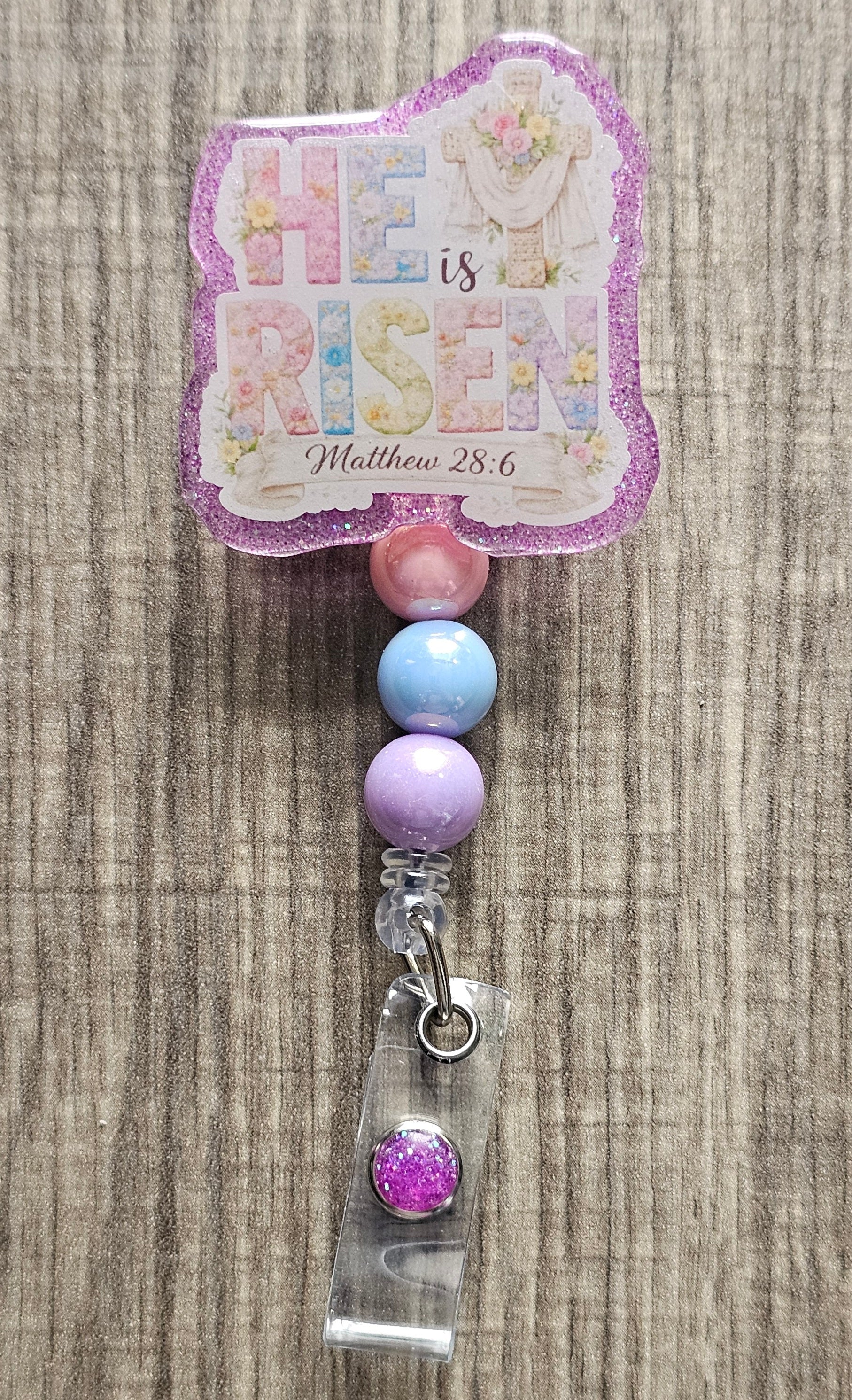 Retractable badge reel featuring "He Is Risen" floral typography with a cross, purple robe, and Matthew 28:6 scripture.