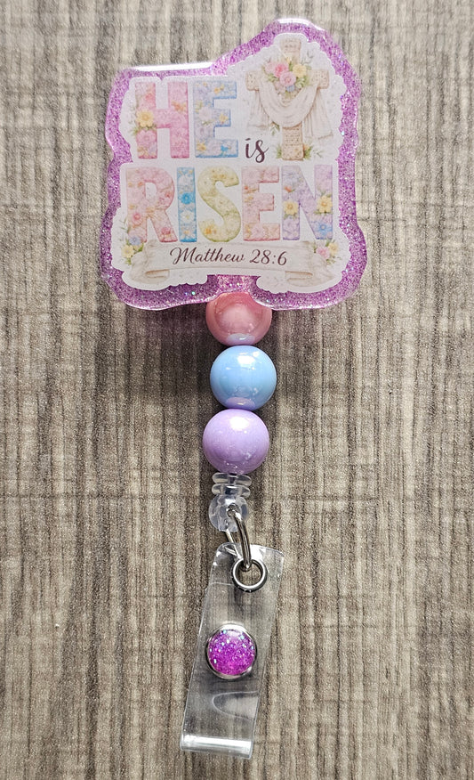 Retractable badge reel featuring "He Is Risen" floral typography with a cross, purple robe, and Matthew 28:6 scripture.
