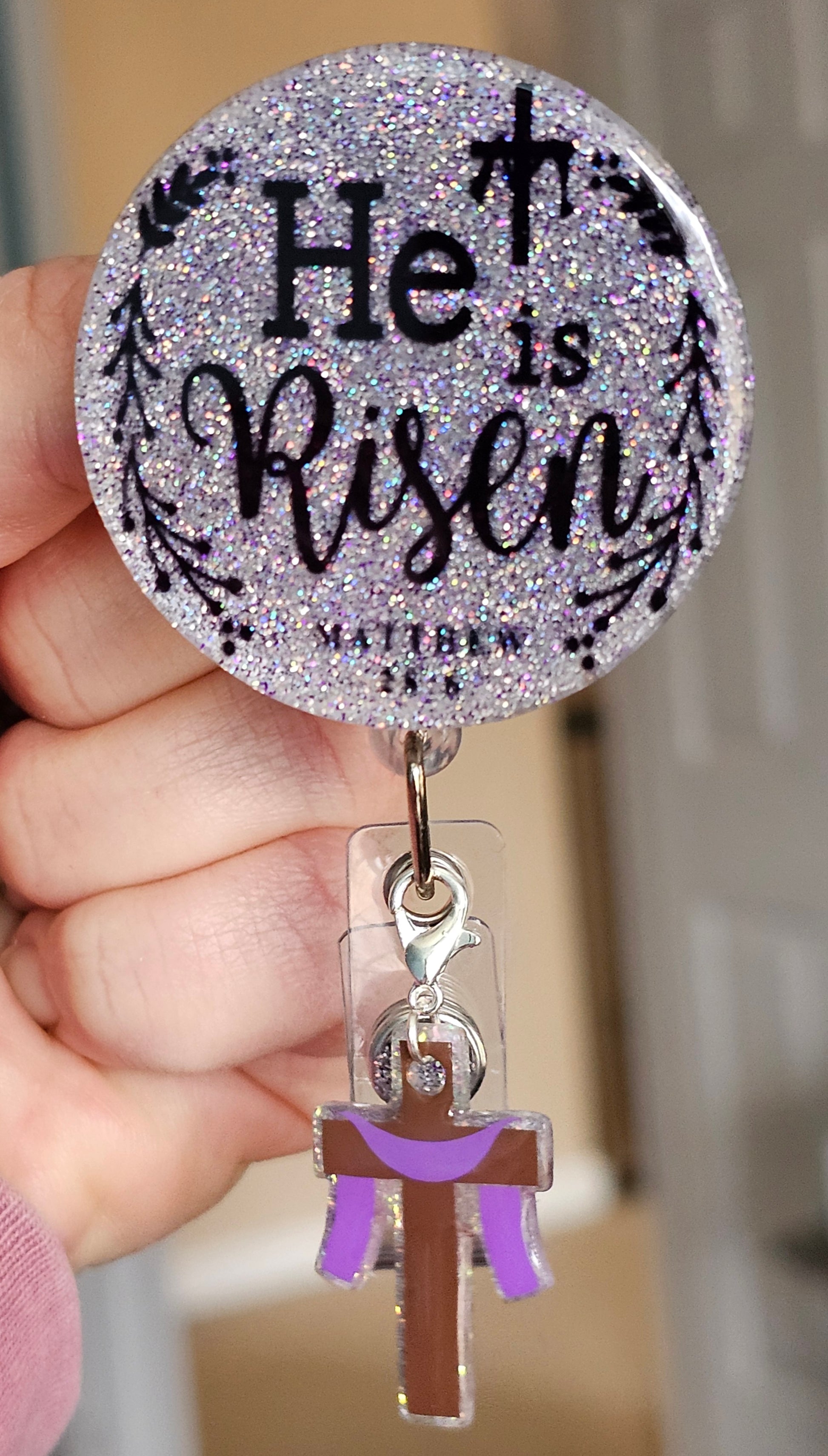 A badge reel featuring "He Is Risen" with Matthew 28:6, a silver glitter background, black text, and a cross clip attachment.