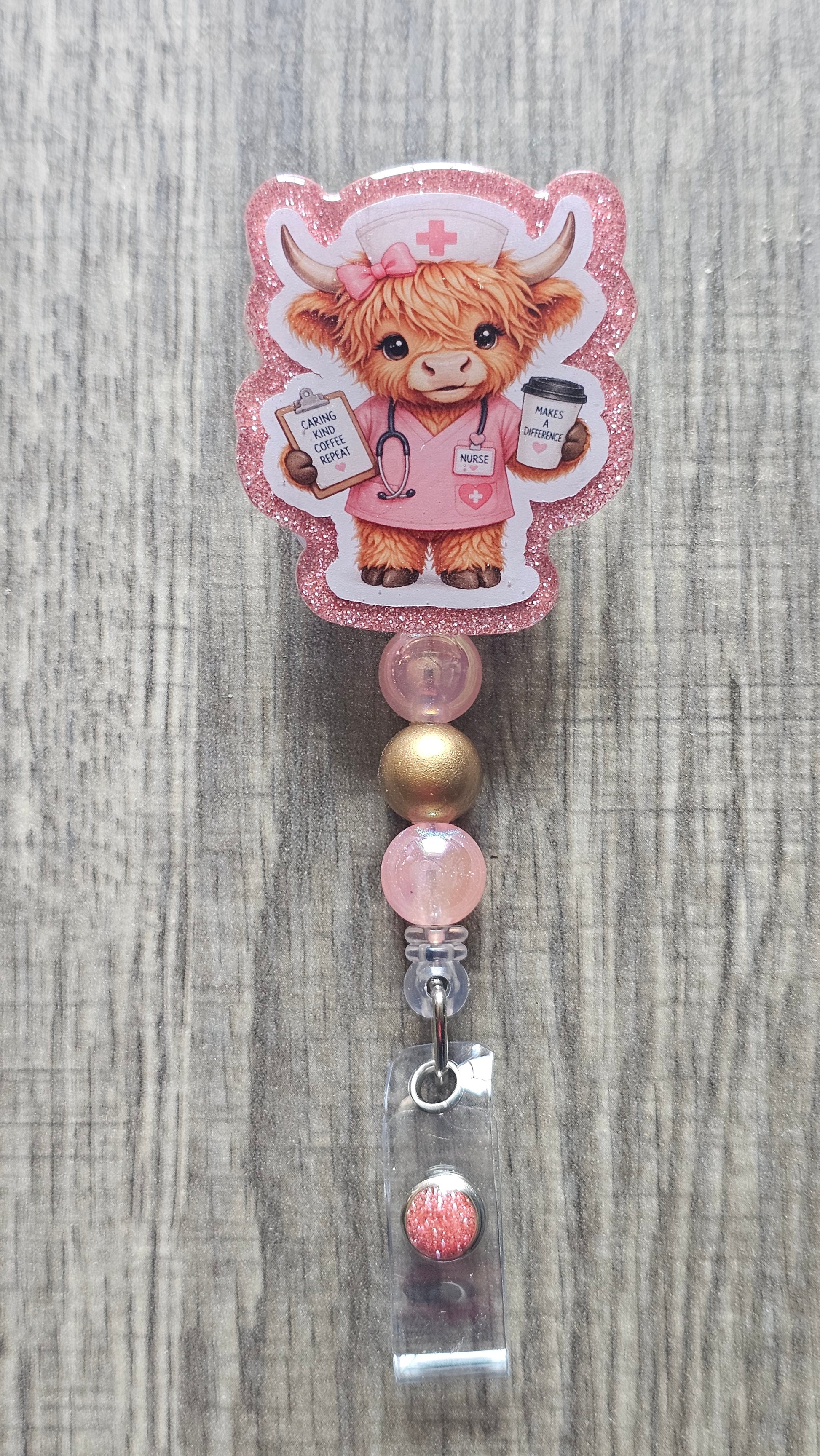 A pink glitter roll-on badge reel featuring a cute cartoon Highland cow dressed as a nurse, with pink and gold beads and a clear plastic clip on a gray wood-textured background.