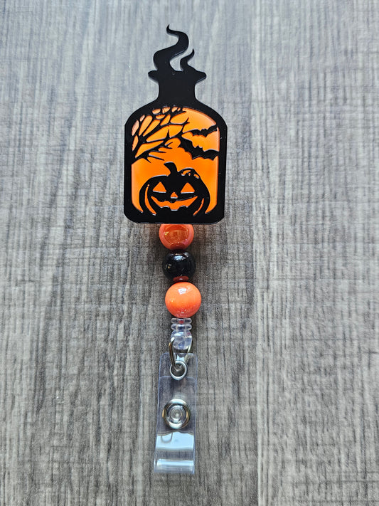Orange glow-in-the-dark potion bottle-shaped badge reel with an eerily smiling jack-o-lantern surrounded by trees and bats, with matching glow-in-the-dark decorative beads.