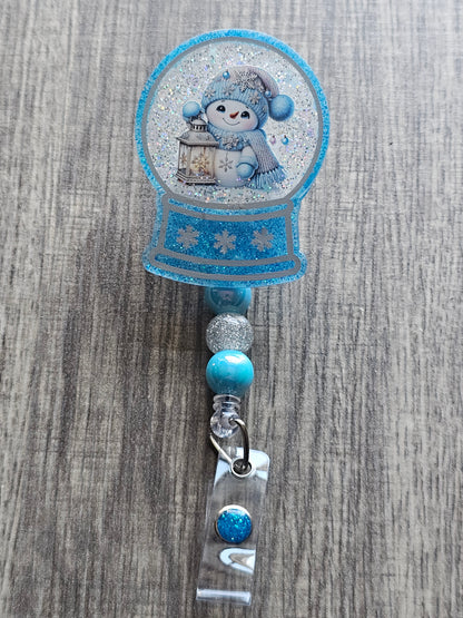 A badge reel with a snow globe design featuring an jolly Snowman in a hat and scarf holding a lantern