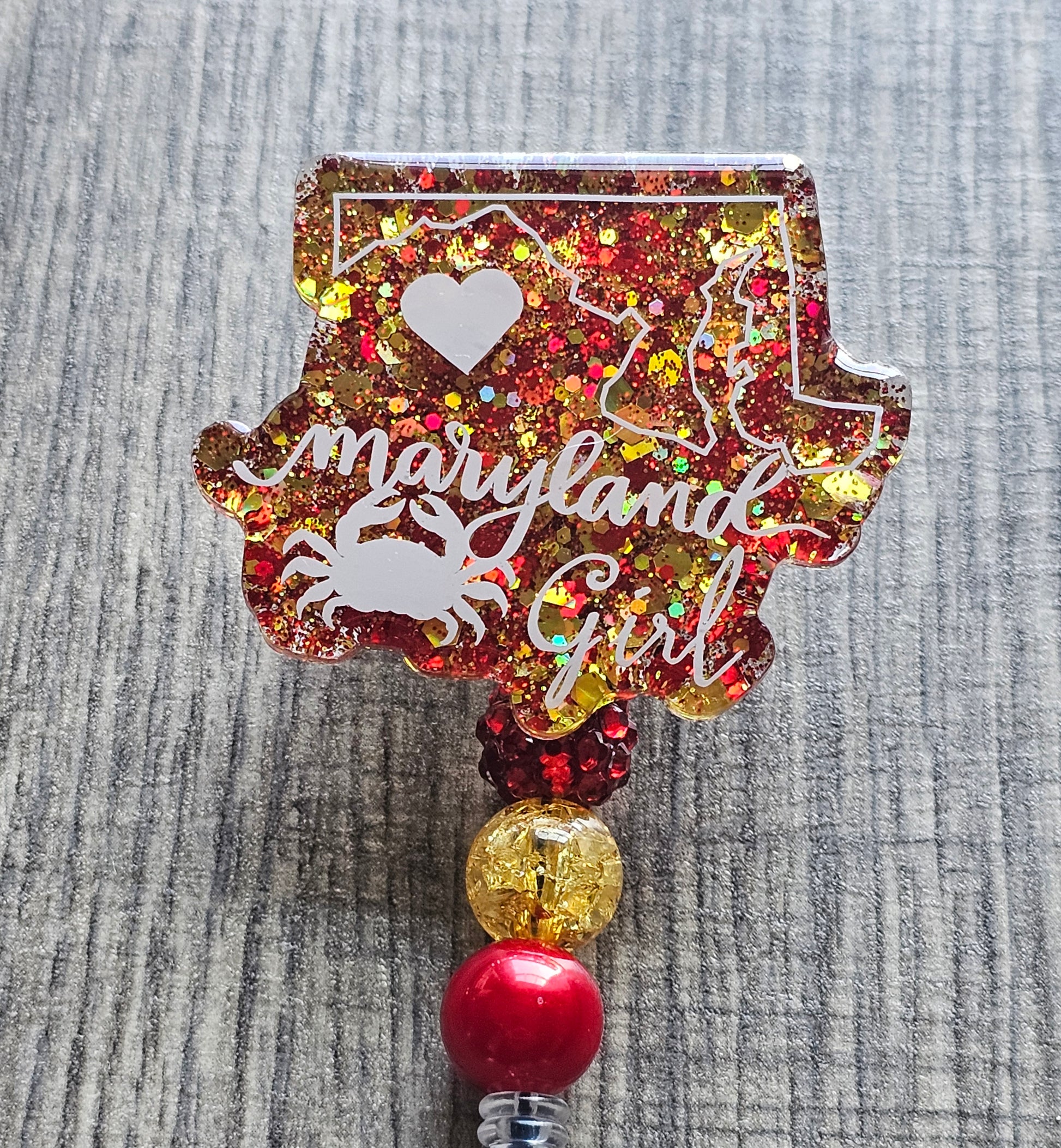 A Badge Reel with a vibrant and stylish design features the shape of the state of Maryland, along with a crab and a heart, all accented with red and yellow glitter to represent the state's colors.