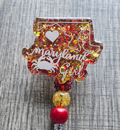 A Badge Reel with a vibrant and stylish design features the shape of the state of Maryland, along with a crab and a heart, all accented with red and yellow glitter to represent the state's colors.