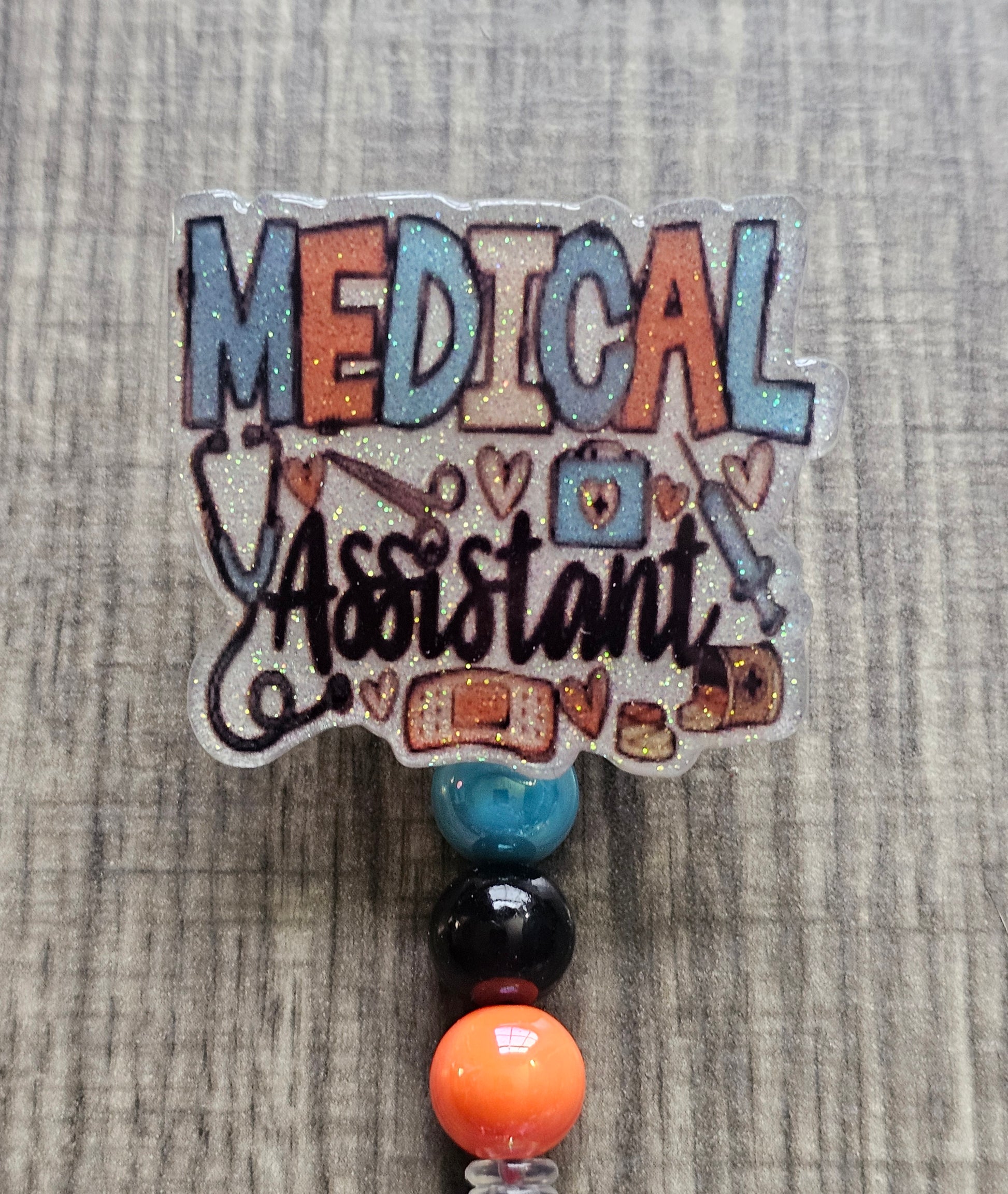 White glitter acrylic badge reel with "Medical Assistant" text, featuring a stethoscope, scissors, first aid kit, syringe, hearts, and medicine, with creatively matching decorative beads.