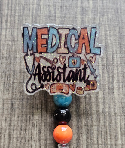 White glitter acrylic badge reel with "Medical Assistant" text, featuring a stethoscope, scissors, first aid kit, syringe, hearts, and medicine, with creatively matching decorative beads.