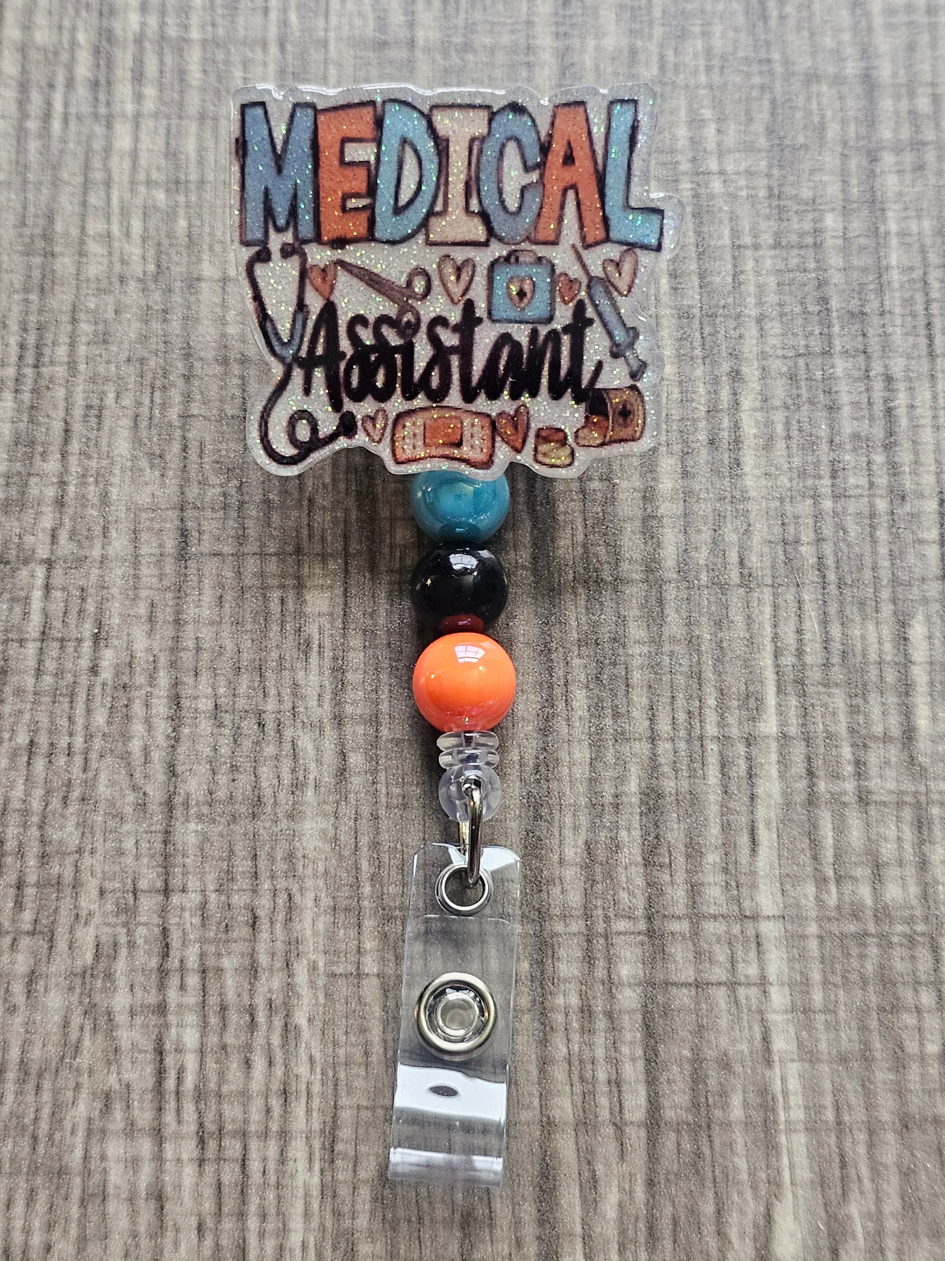 White glitter acrylic badge reel with "Medical Assistant" text, featuring a stethoscope, scissors, first aid kit, syringe, hearts, and medicine, with creatively matching decorative beads.