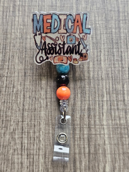 White glitter acrylic badge reel with "Medical Assistant" text, featuring a stethoscope, scissors, first aid kit, syringe, hearts, and medicine, with creatively matching decorative beads.