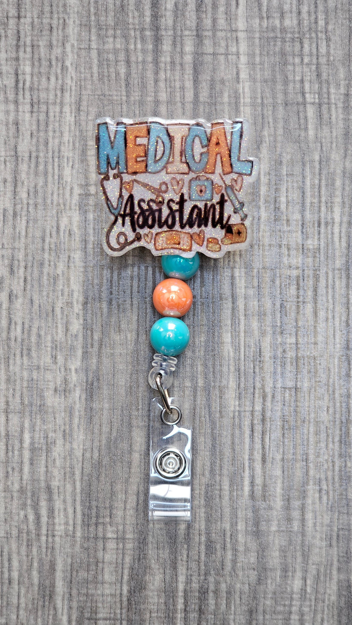 White glitter acrylic badge reel with "Medical Assistant" text, featuring a stethoscope, scissors, first aid kit, syringe, hearts, and medicine, with creatively matching decorative beads.