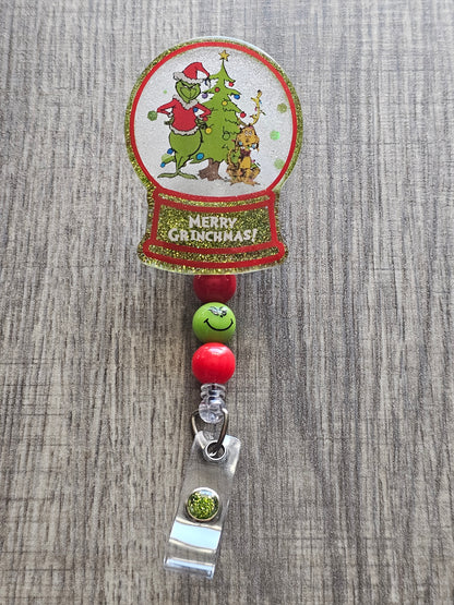 A badge reel with a snow globe design featuring the grinch standing beside a Christmas tree in a Christmas sweater and hat along his dog whose wrapped with Christmas lights