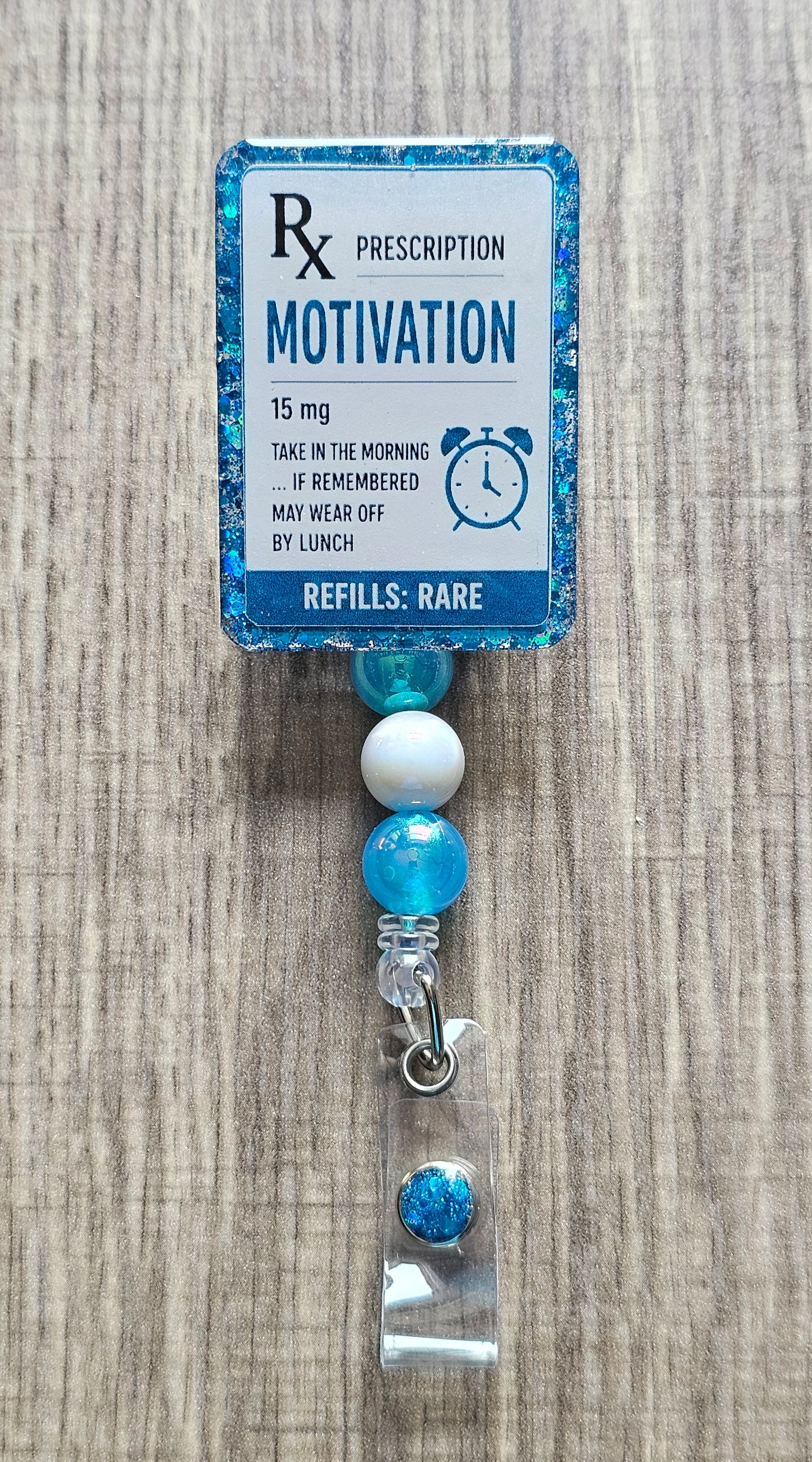 A novelty prescription-style badge reel with a blue glitter border and a printed label reading 'Rx Prescription Motivation 15 mg' on a wood surface.