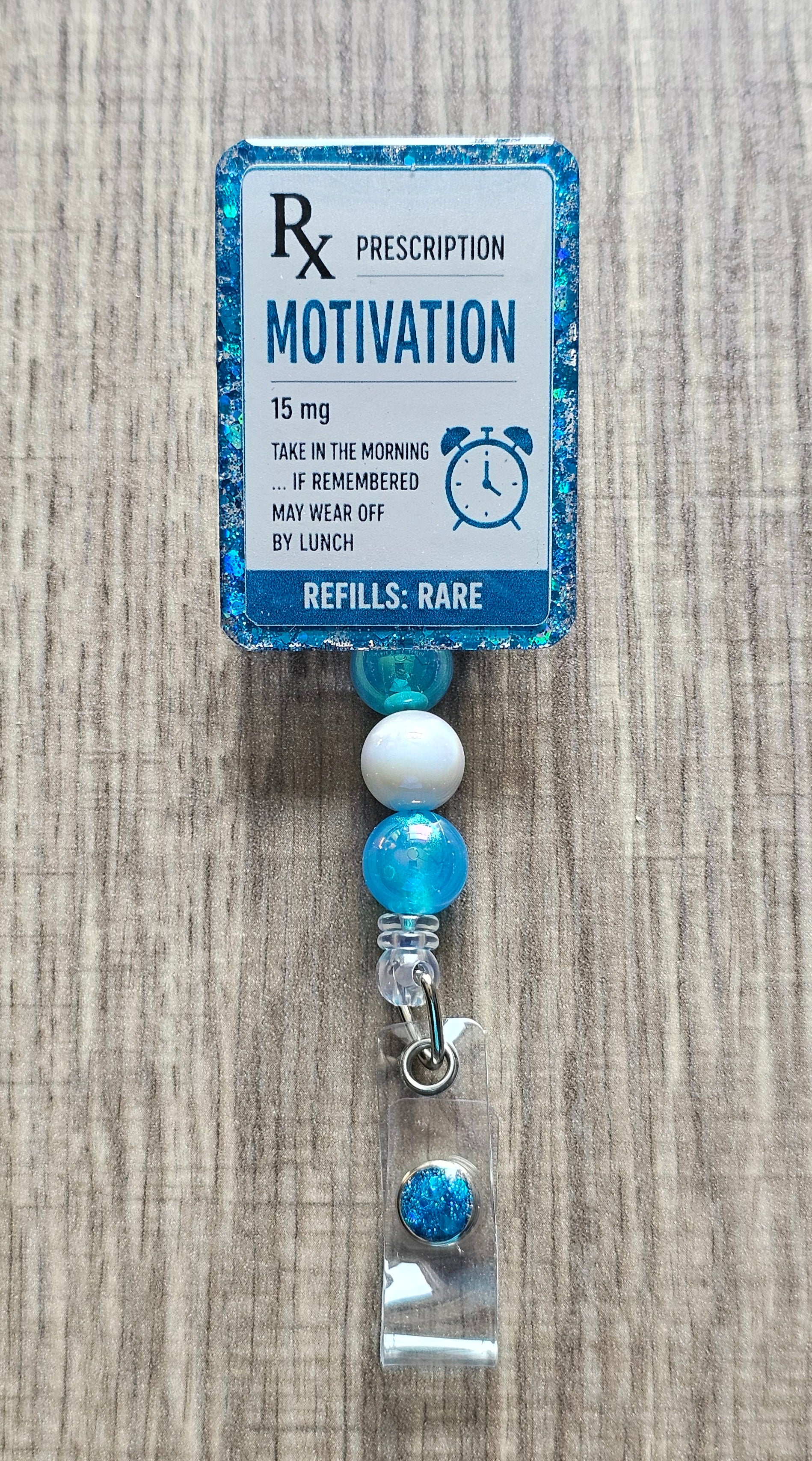 A novelty prescription-style badge reel with a blue glitter border and a printed label reading 'Rx Prescription Motivation 15 mg' on a wood surface.