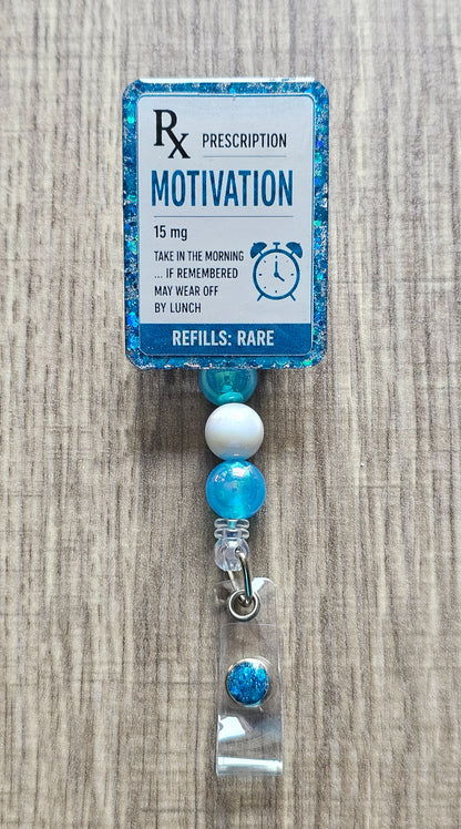 A novelty prescription-style badge reel with a blue glitter border and a printed label reading 'Rx Prescription Motivation 15 mg' on a wood surface.