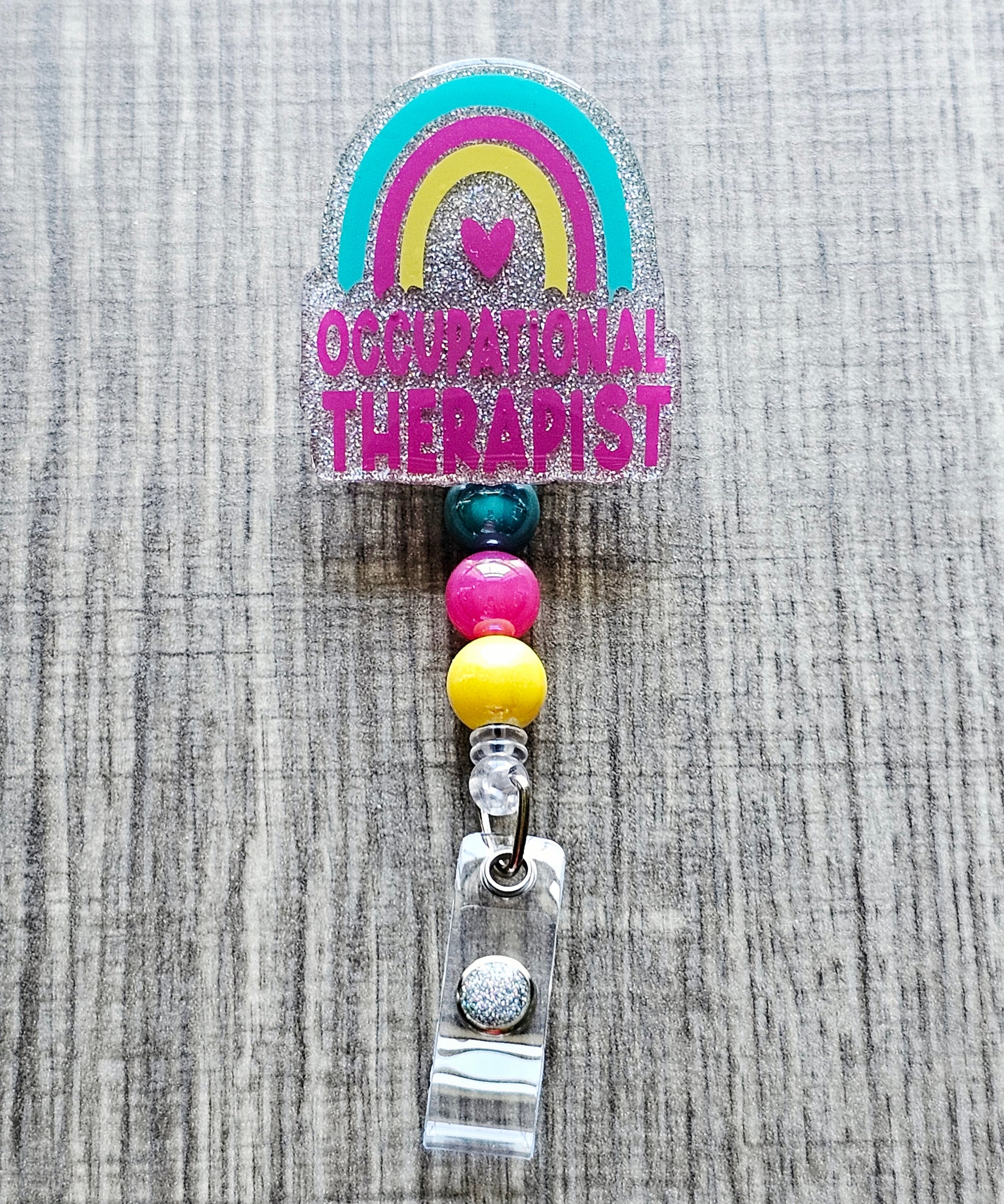 Glitter occupational therapist badge reel with a rainbow and heart design.
