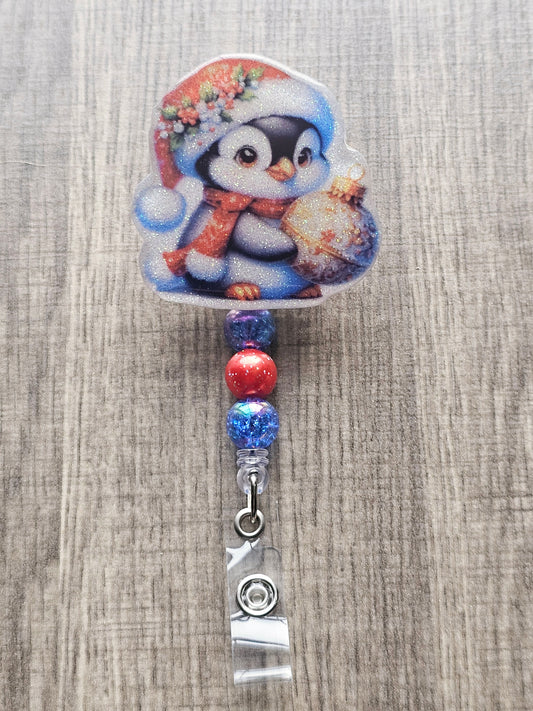 A badge reel featuring an adorable penguin dressed in a cozy Christmas hat and scarf, holding a bright and cheerful Christmas ornament.