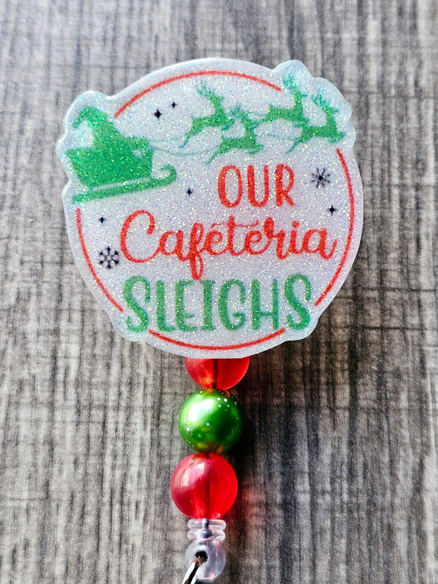 A badge reel featuring Santa flying across the sky in his sleigh with his reindeer, this badge reel is adorned with vibrant Christmas-themed colors along with the words Our Cafeteria Sleighs