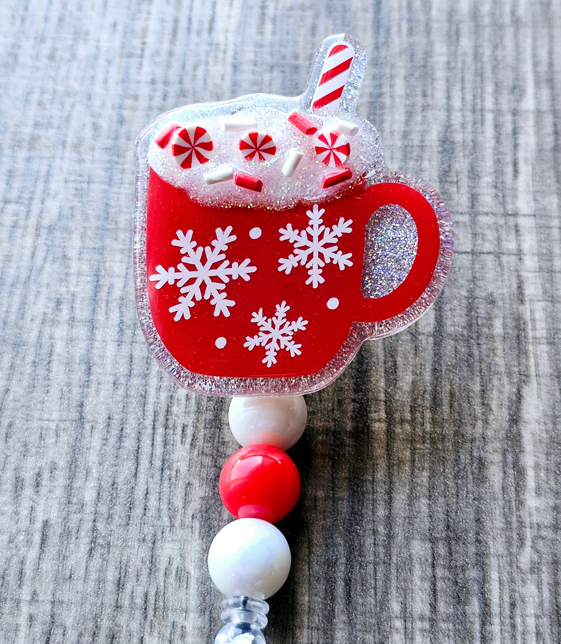 A festive badge reel shaped like a red mug, adorned with a delicate snowflake design. Topped with playful peppermint candy.