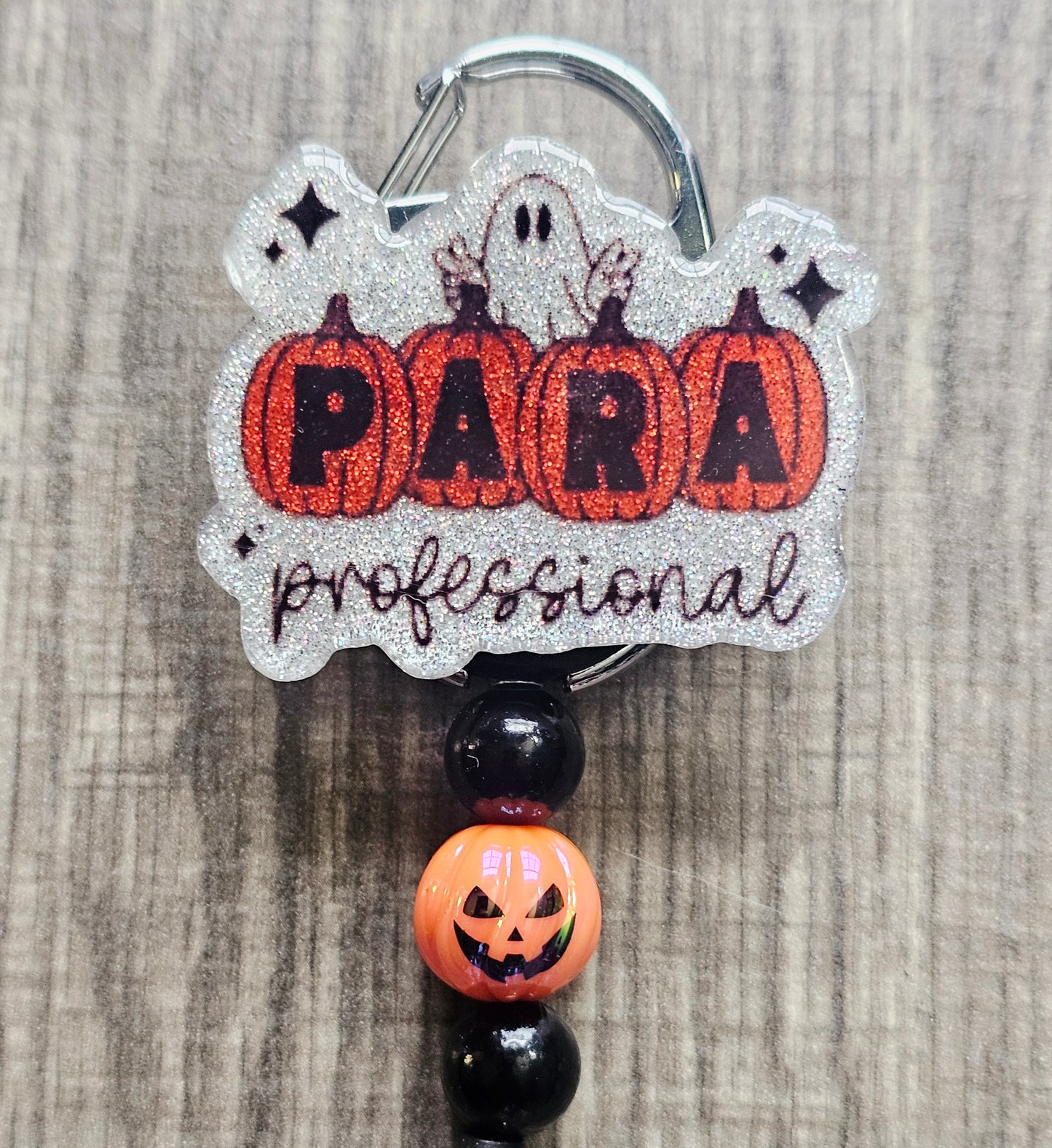 White glitter acrylic badge reel with pumpkins spelling "Para," "Professional" written below, a ghost behind the pumpkins, and matching beads with a central jack-o-lantern bead.