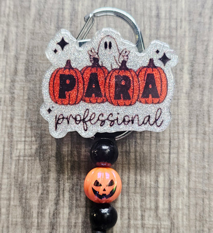 White glitter acrylic badge reel with pumpkins spelling "Para," "Professional" written below, a ghost behind the pumpkins, and matching beads with a central jack-o-lantern bead.