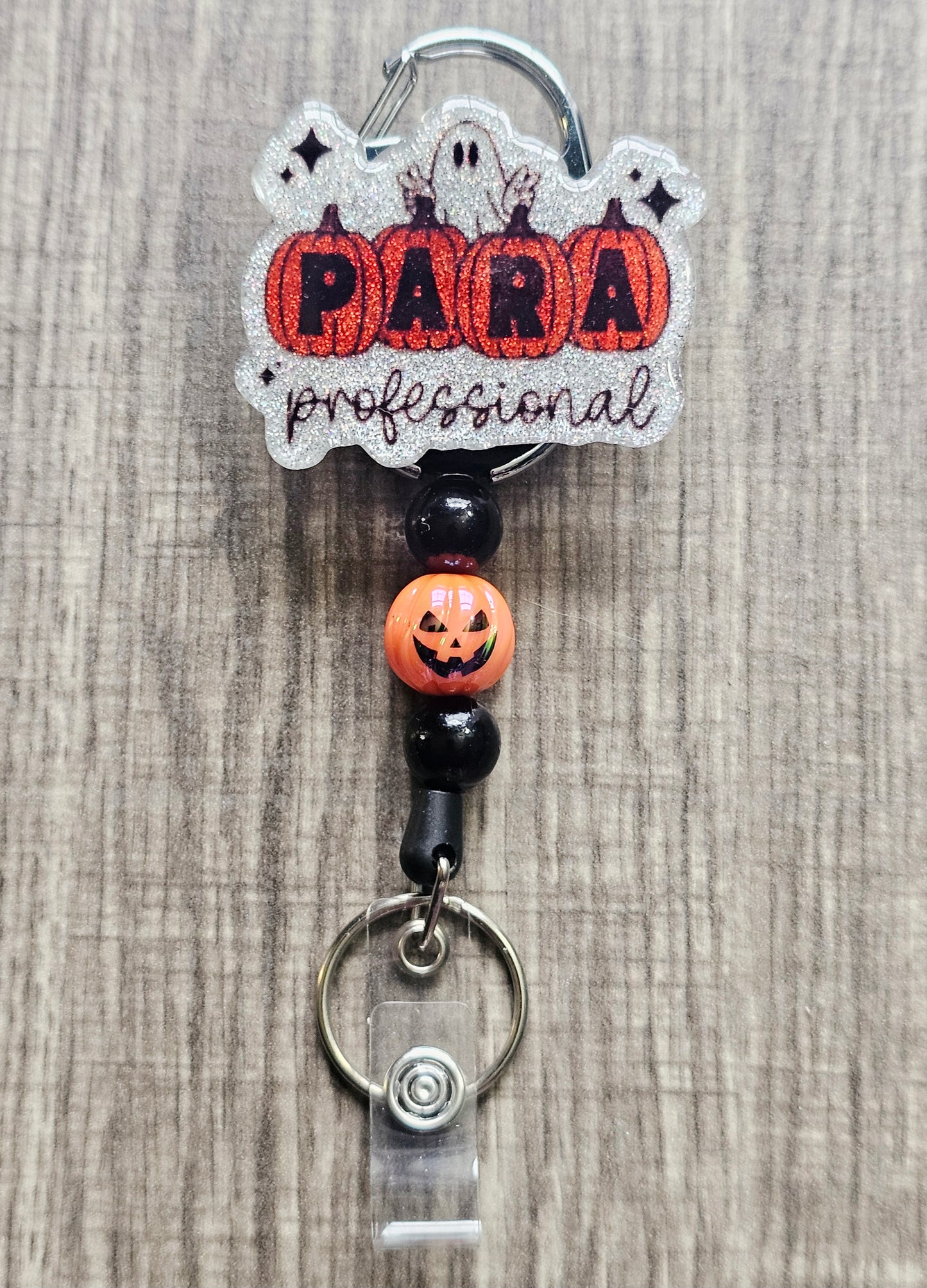 White glitter acrylic badge reel with pumpkins spelling "Para," "Professional" written below, a ghost behind the pumpkins, and matching beads with a central jack-o-lantern bead.