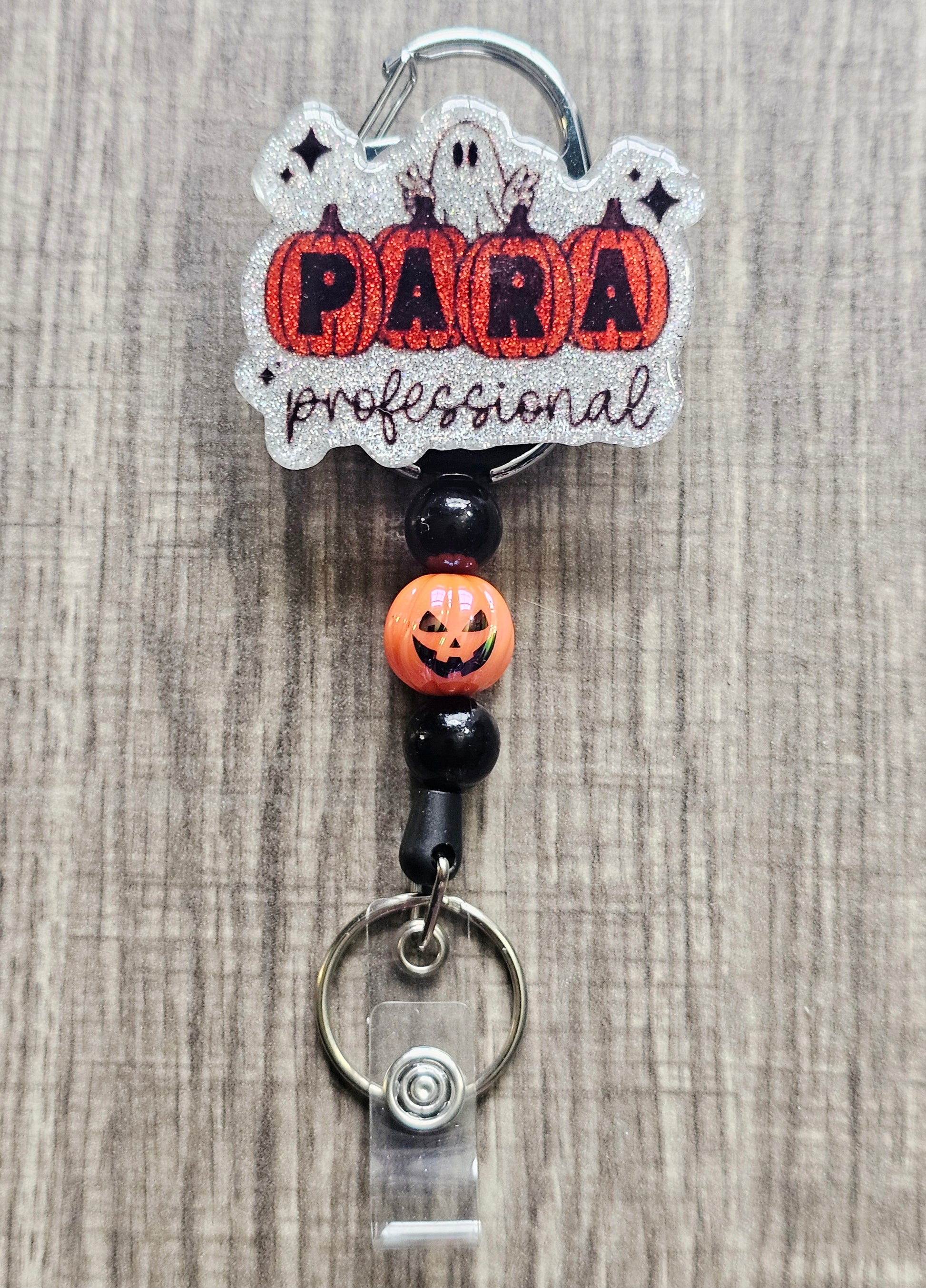 White glitter acrylic badge reel with pumpkins spelling "Para," "Professional" written below, a ghost behind the pumpkins, and matching beads with a central jack-o-lantern bead.