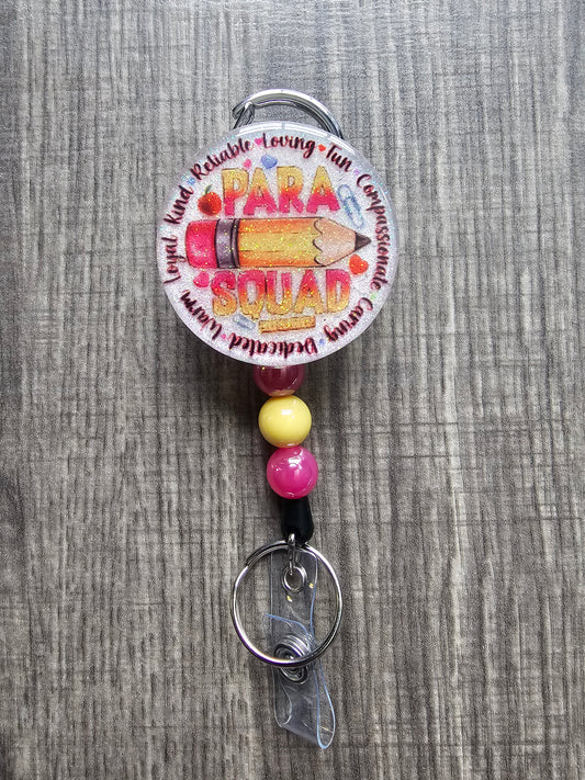 White glitter acrylic badge reel with a pencil, apple, ruler, paper clip, and "Para Squad" text surrounded by traits like "Loyal," "Kind," "Reliable," and "Dedicated."