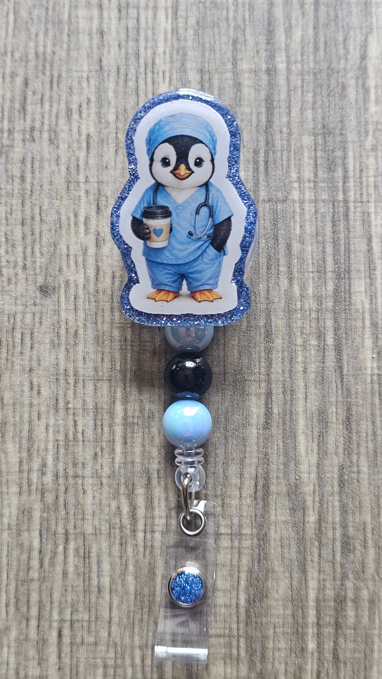 A penguin-shaped badge reel or clip featuring a cartoon penguin dressed in blue medical scrubs, standing on a light wood surface. The reel has a blue glitter border, and the hanging section includes dark blue, light blue, and clear beads with a silver clasp.