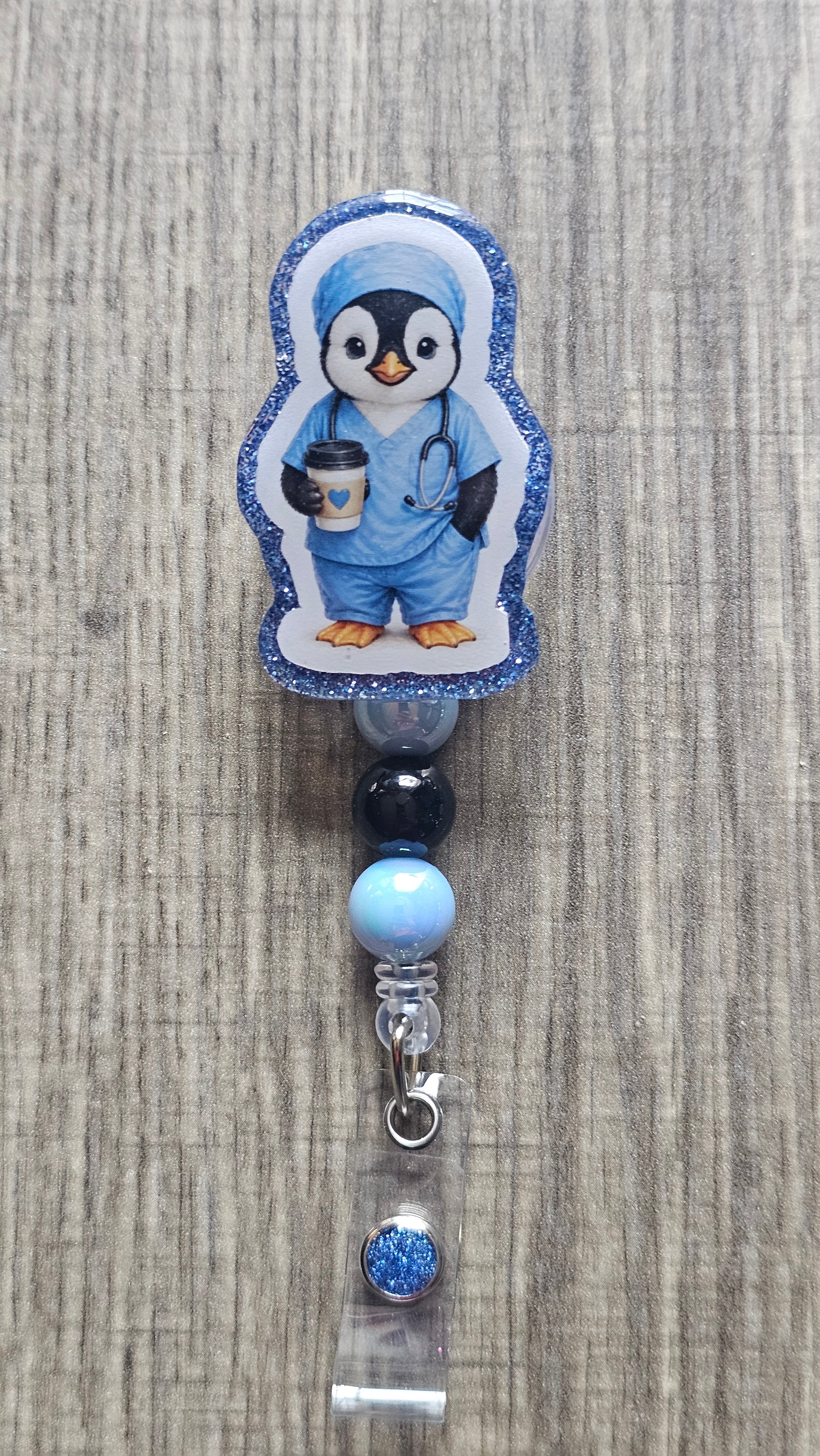A penguin-shaped badge reel or clip featuring a cartoon penguin dressed in blue medical scrubs, standing on a light wood surface. The reel has a blue glitter border, and the hanging section includes dark blue, light blue, and clear beads with a silver clasp.