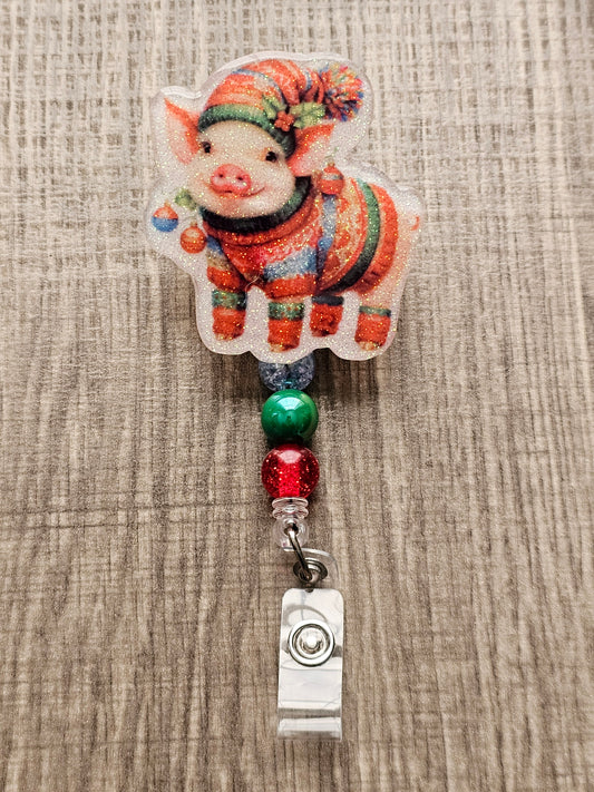 A badge reel that displays an adorable piglet in a festive sweater and hat adorned with Christmas ornaments and a charming smile 