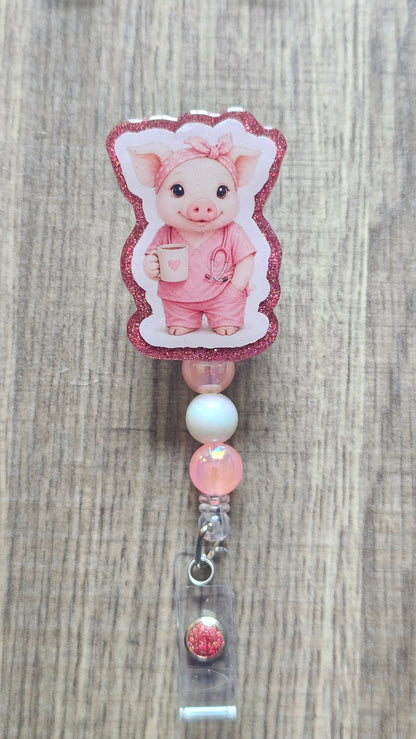 A pink pig-shaped retractable badge reel with a glittery pink border, featuring a cartoon pig character in pink scrubs. The reel is shown attached to a clear plastic clip with a pink and white bead spacer, set against a light brown wood grain background.