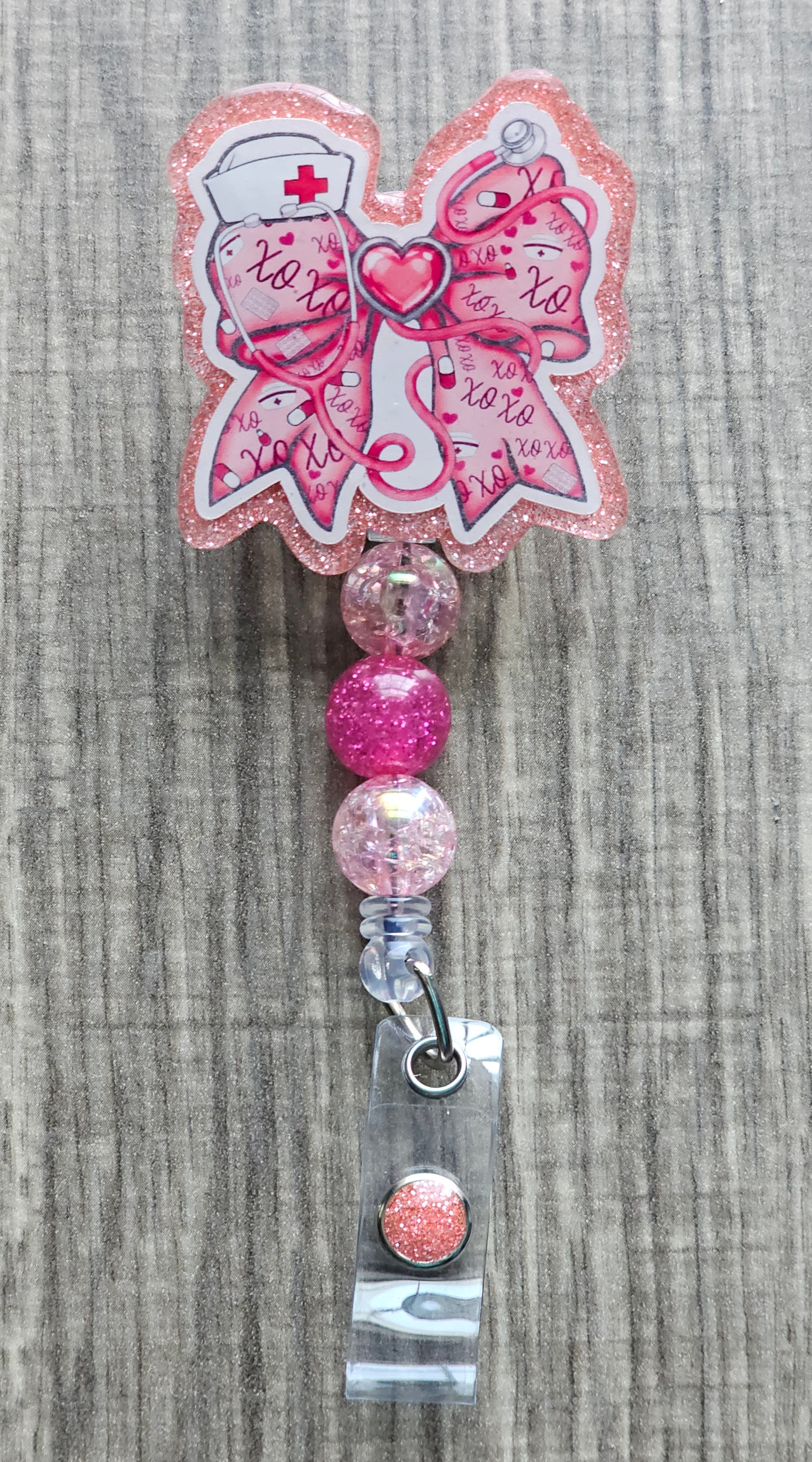 Pink glitter retractable nurse badge reel with a bow-shaped acrylic top featuring medical illustrations, 'xoxo' text, and pink beaded accents. Attaches via a 360-degree swivel alligator clip.