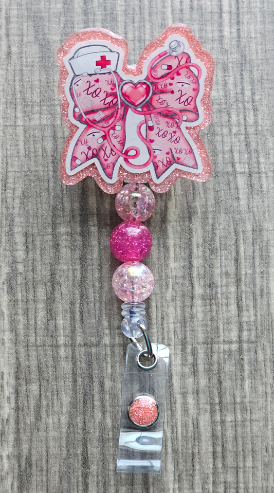 Pink glitter retractable nurse badge reel with a bow-shaped acrylic top featuring medical illustrations, 'xoxo' text, and pink beaded accents. Attaches via a 360-degree swivel alligator clip.