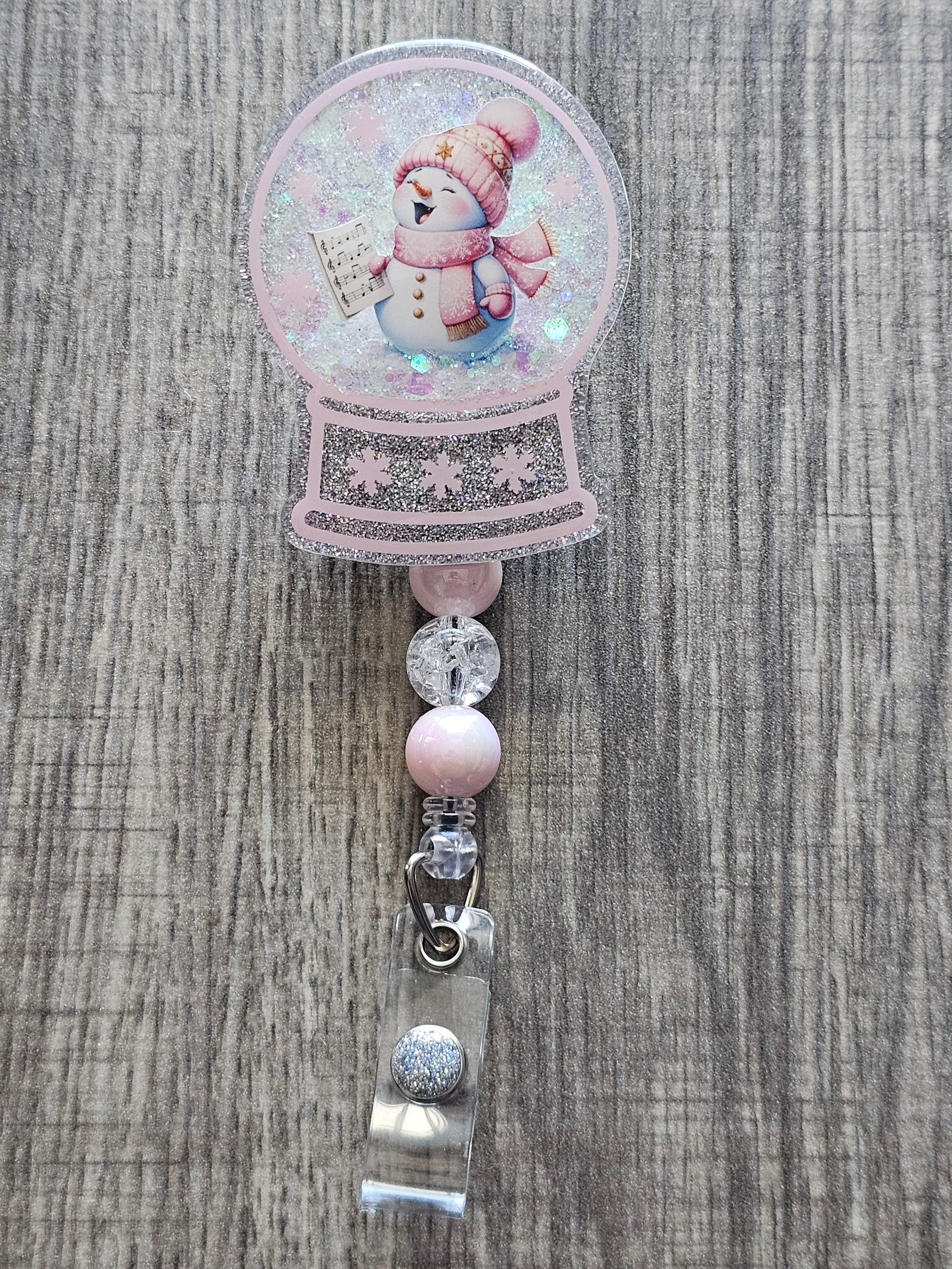 A badge reel with a snow globe design featuring an adorable snowman adorned with a pink hat and scarf holding a sheet of music singing.