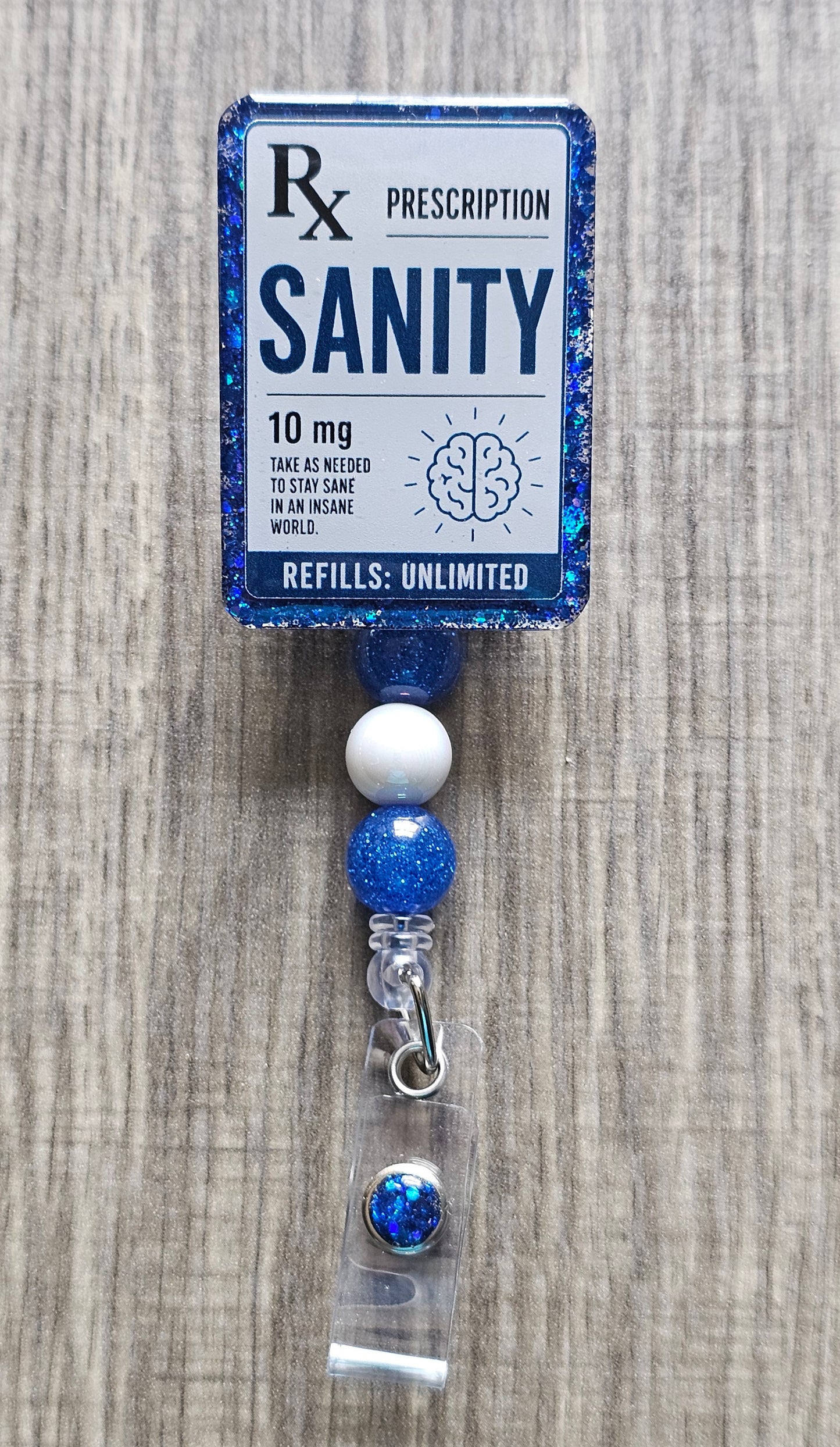 A blue glitter prescription-style card holder with the word 'SANITY' and Rx symbol, attached to blue and white beads and a clear clip on a gray wood-textured background.