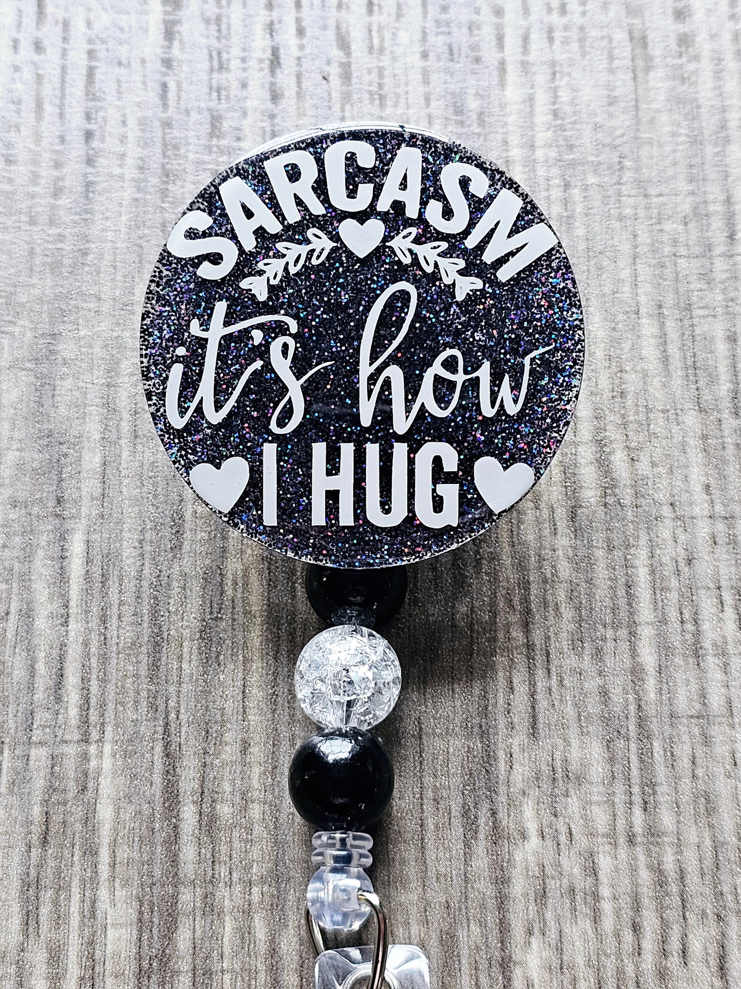 A badge reel with a glittery black background featuring the phrase "Sarcasm: It’s How I Hug" in bold white font.