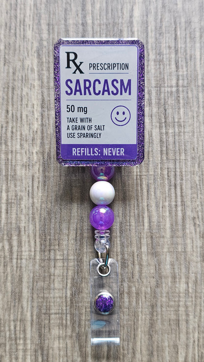 A novelty prescription-style beaded accessory with a printed card reading 'SARCASM 50 mg' and a dangling clear case containing a purple glitter bead, shown on a wood surface.