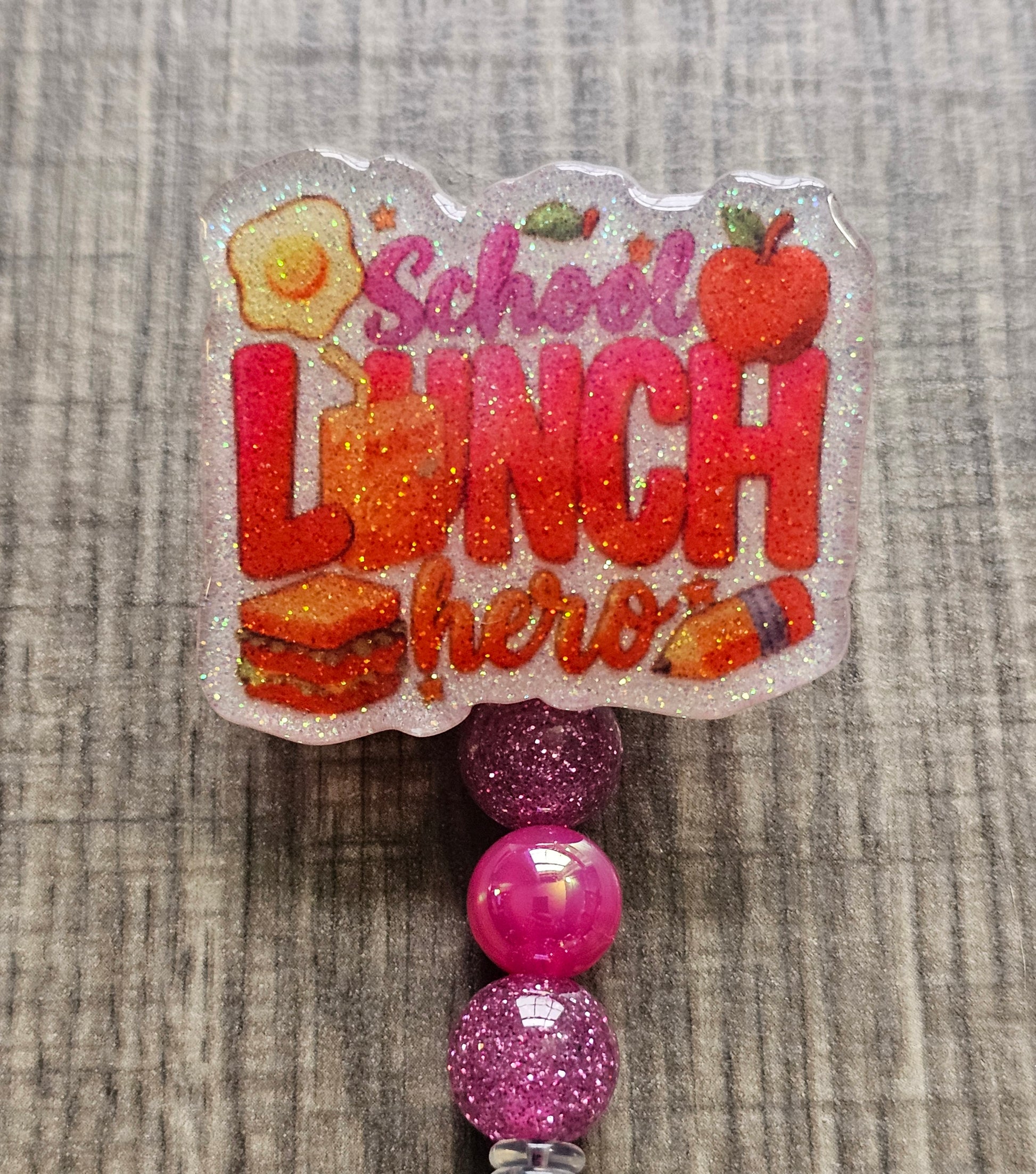 White glitter acrylic badge reel with "School Lunch Hero" text, creatively incorporating a juice box, sandwich, pencil, apple, and egg, with matching decorative beads.