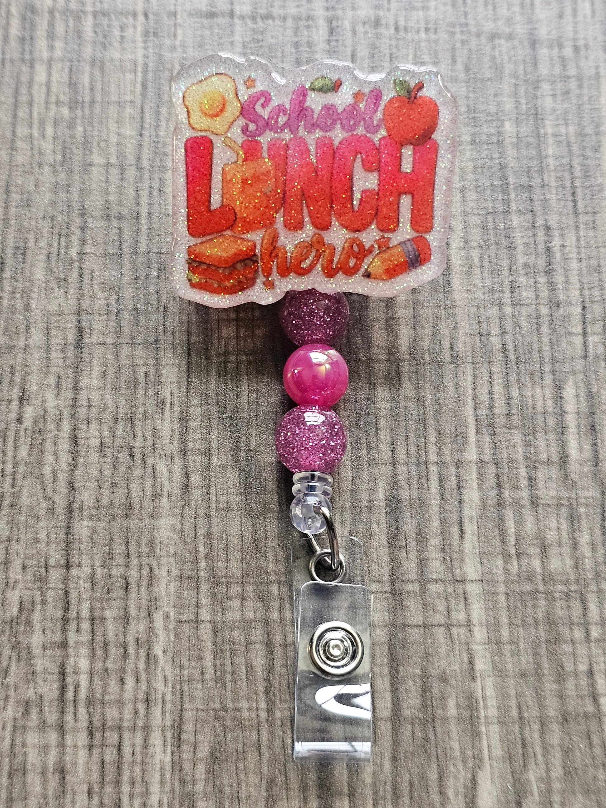 White glitter acrylic badge reel with "School Lunch Hero" text, creatively incorporating a juice box, sandwich, pencil, apple, and egg, with matching decorative beads.