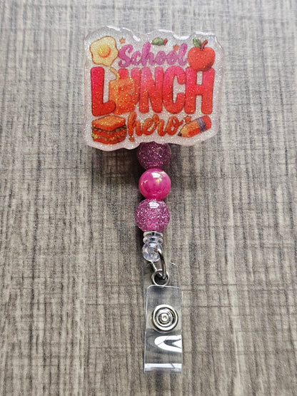 White glitter acrylic badge reel with "School Lunch Hero" text, creatively incorporating a juice box, sandwich, pencil, apple, and egg, with matching decorative beads.