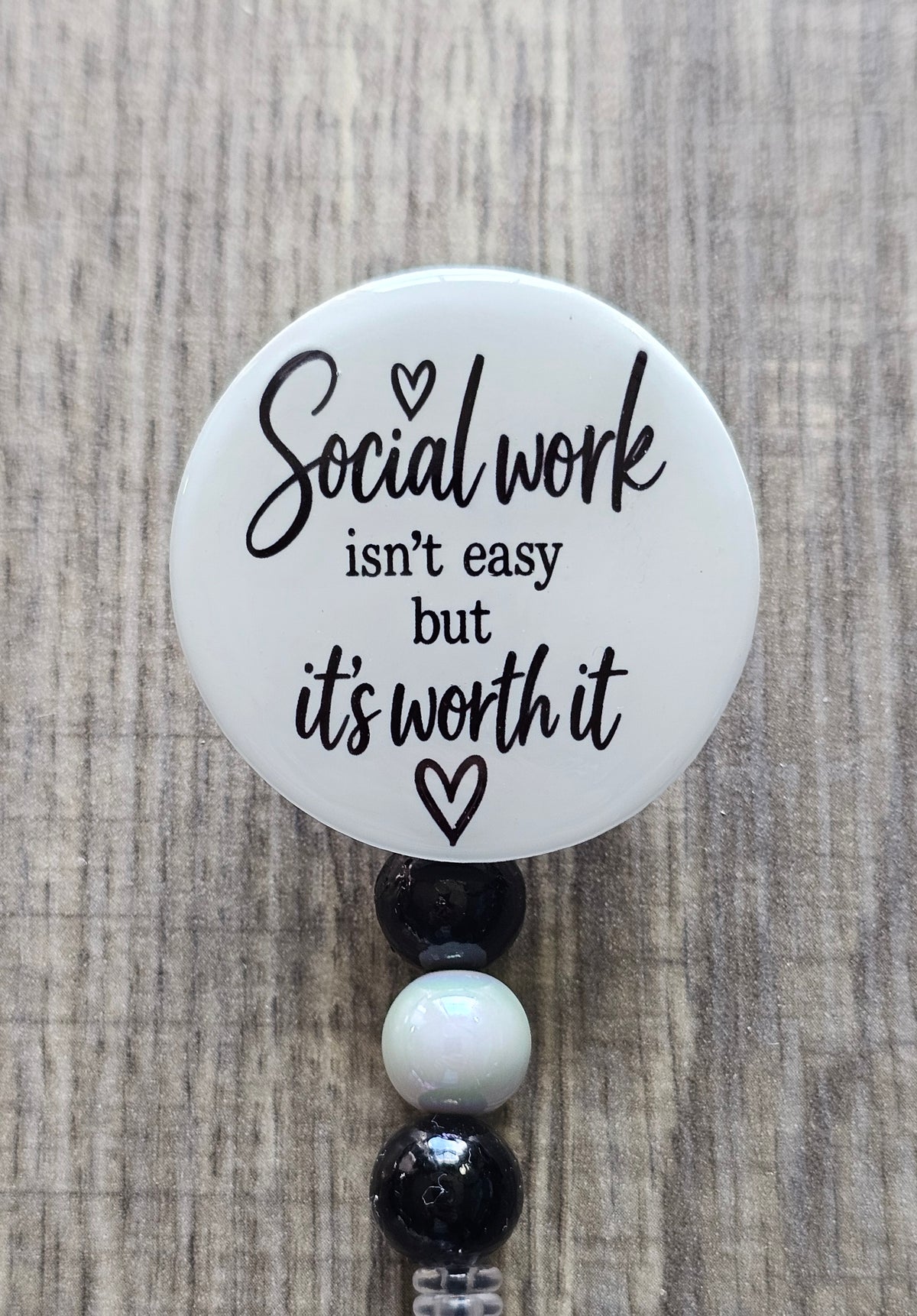 Retractable social worker beaded badge reel with a white faceplate, black text reading "Social Work isn't easy but it's worth it" and a small heart, adorned with coordinating black and white beads.