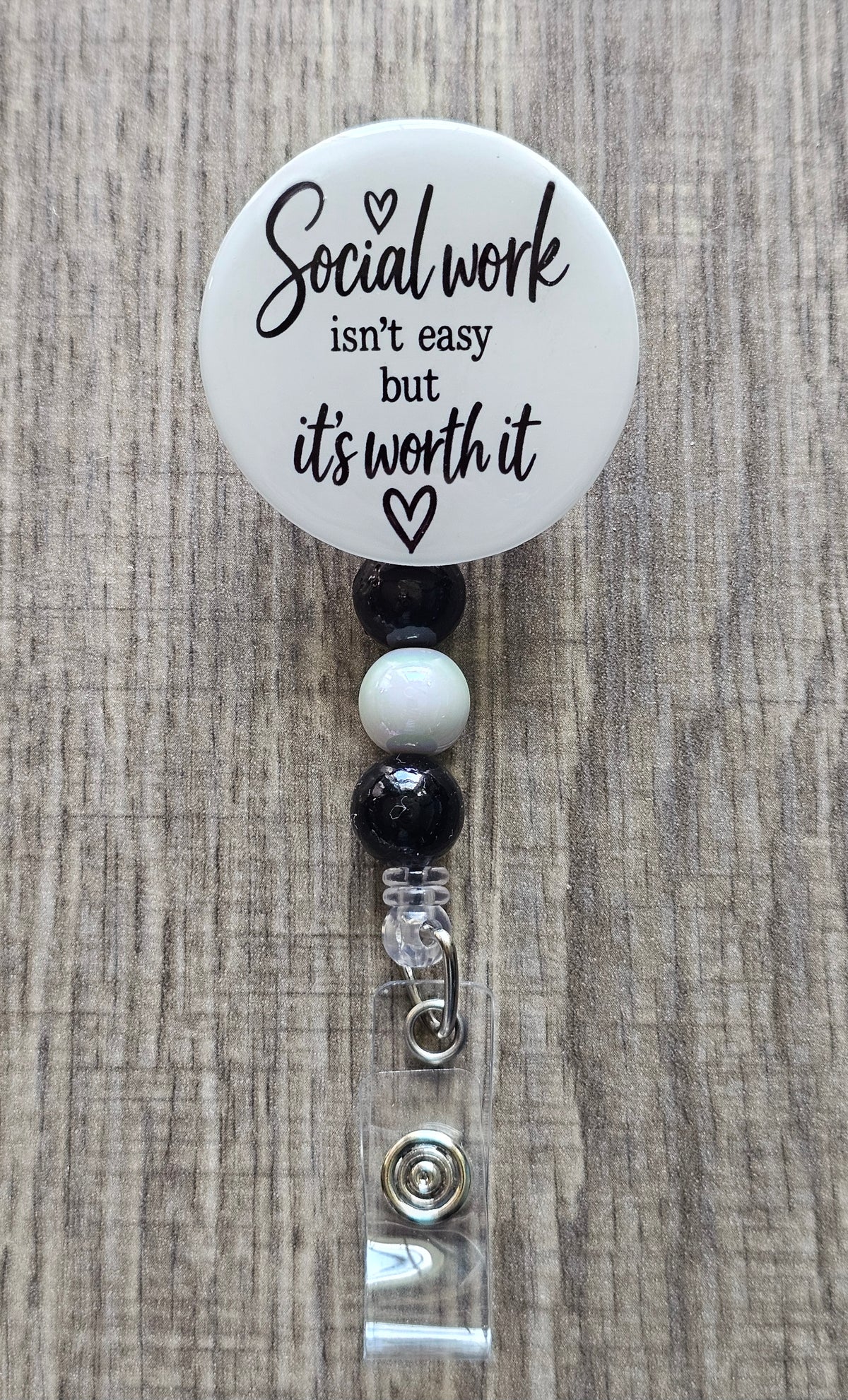 Retractable social worker beaded badge reel with a white faceplate, black text reading "Social Work isn't easy but it's worth it" and a small heart, adorned with coordinating black and white beads.