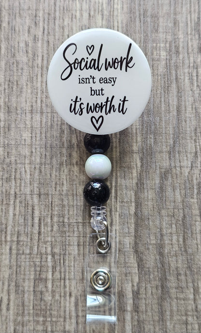 Retractable social worker beaded badge reel with a white faceplate, black text reading "Social Work isn't easy but it's worth it" and a small heart, adorned with coordinating black and white beads.