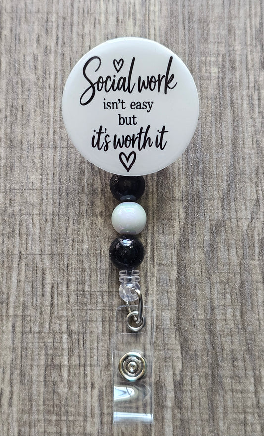 Retractable social worker beaded badge reel with a white faceplate, black text reading "Social Work isn't easy but it's worth it" and a small heart, adorned with coordinating black and white beads.