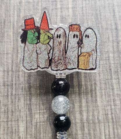 Badge reel depicting five figures in white ghost costumes, two wearing witch masks, and one holding a trick-or-treat bag, with matching decorative beads.