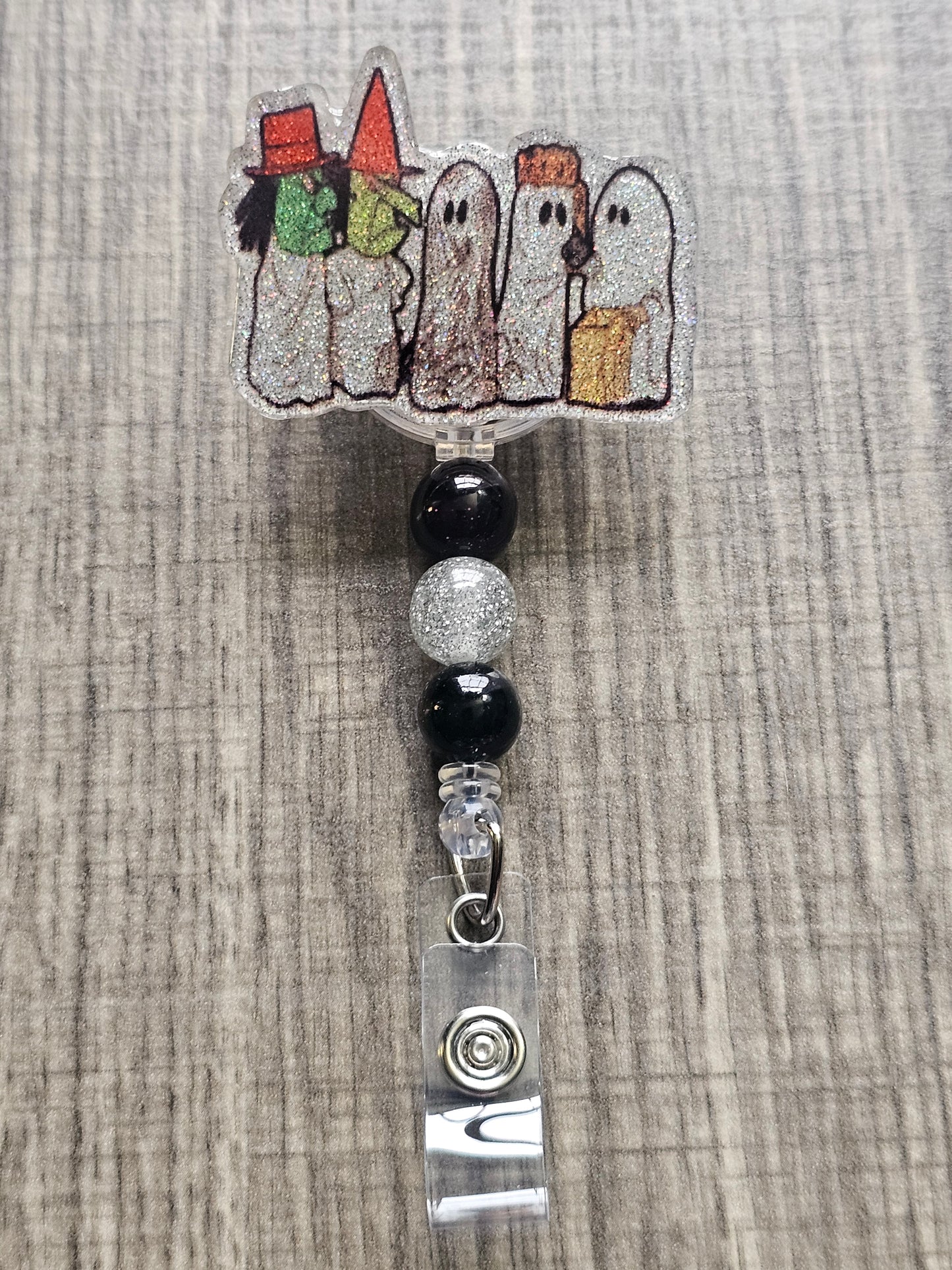 Badge reel depicting five figures in white ghost costumes, two wearing witch masks, and one holding a trick-or-treat bag, with matching decorative beads.